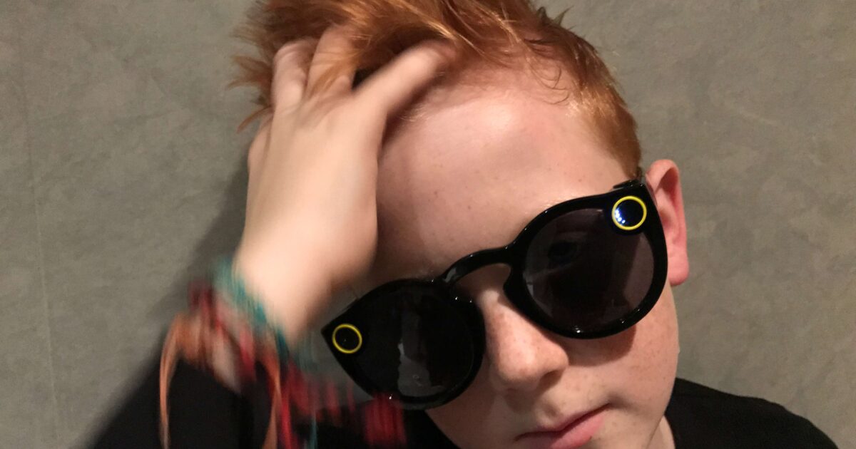 En ung pojke med rött hår som bär svarta eleganta solglasögon rör vid håret med ena handen. Han är klädd i en svart topp och har färgglada armband på handleden, stående mot en enfärgad grå bakgrund.