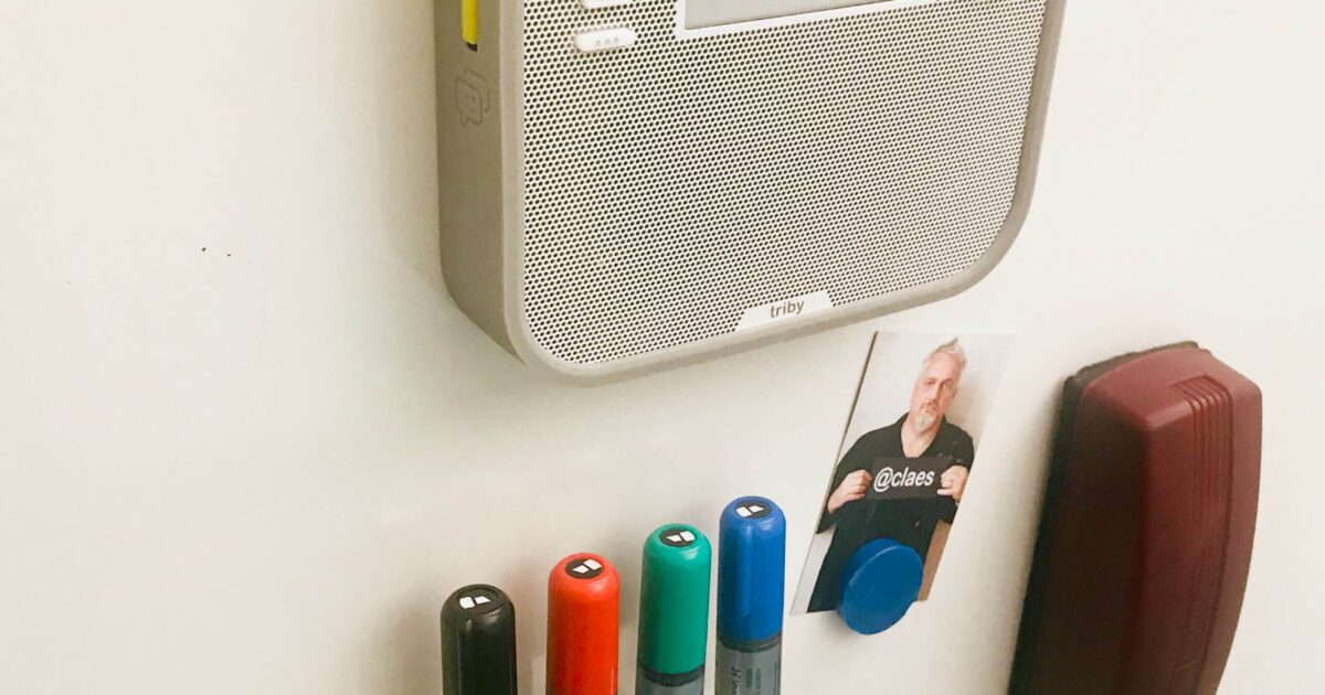 En whiteboardtavla med en monterad radio, fyrfärgade markörer i en hållare, ett litet foto av en person med en bricka och ett brunt suddgummi fäst på tavlan.