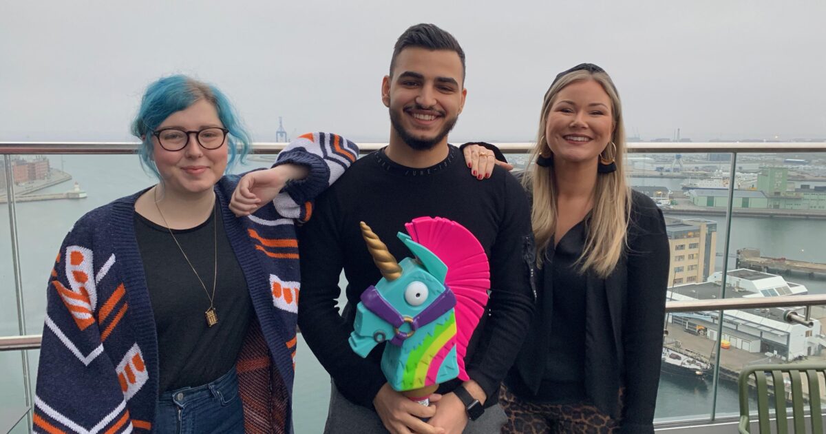 Tre personer står på ett tak vid ett räcke med en stad och en hamn i bakgrunden. Personen i mitten håller en färgglad leksaksenhörning. Alla tre ler mot kameran.