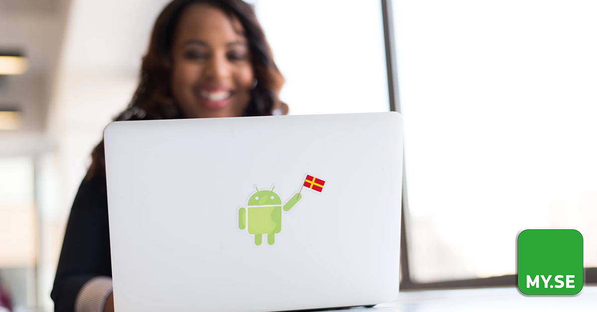 En leende kvinna sitter vid ett skrivbord och använder en bärbar dator med ett Android-klistermärke med en dansk flagga. MY.SE-logotypen syns i det nedre högra hörnet. Solljus strömmar in genom stora fönster i bakgrunden.