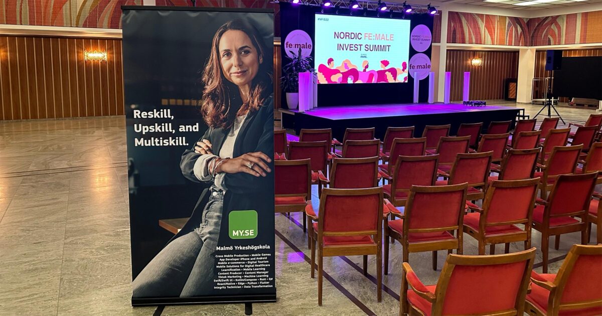 En konferenssal med rader av röda stolar mittemot en scen och en skärm där det står Nordic Female Invest Summit. I förgrunden syns en banderoll med en leende kvinna och text som förespråkar omskolning och kompetensutveckling.