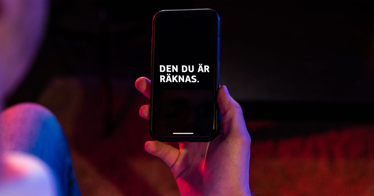 En person håller en smartphone som visar den svenska texten DEN DU ÄR RÄKNAS i vitt på en svart skärm. Bakgrunden är svagt upplyst med rosa och lila toner.