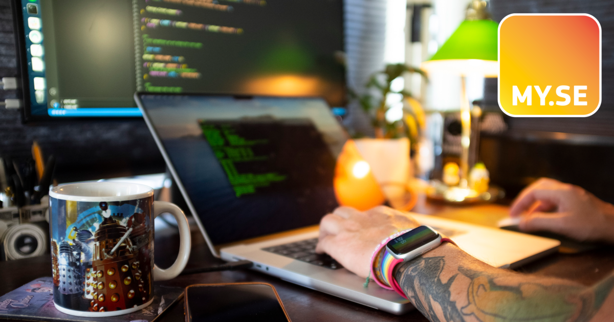 En person med tatuering och smartklocka skriver på en bärbar dator vid ett skrivbord med kodning på skärmen, en Dalek-mugg, en telefon och ett ljus. MY.SE-logotypen visas i det övre högra hörnet.