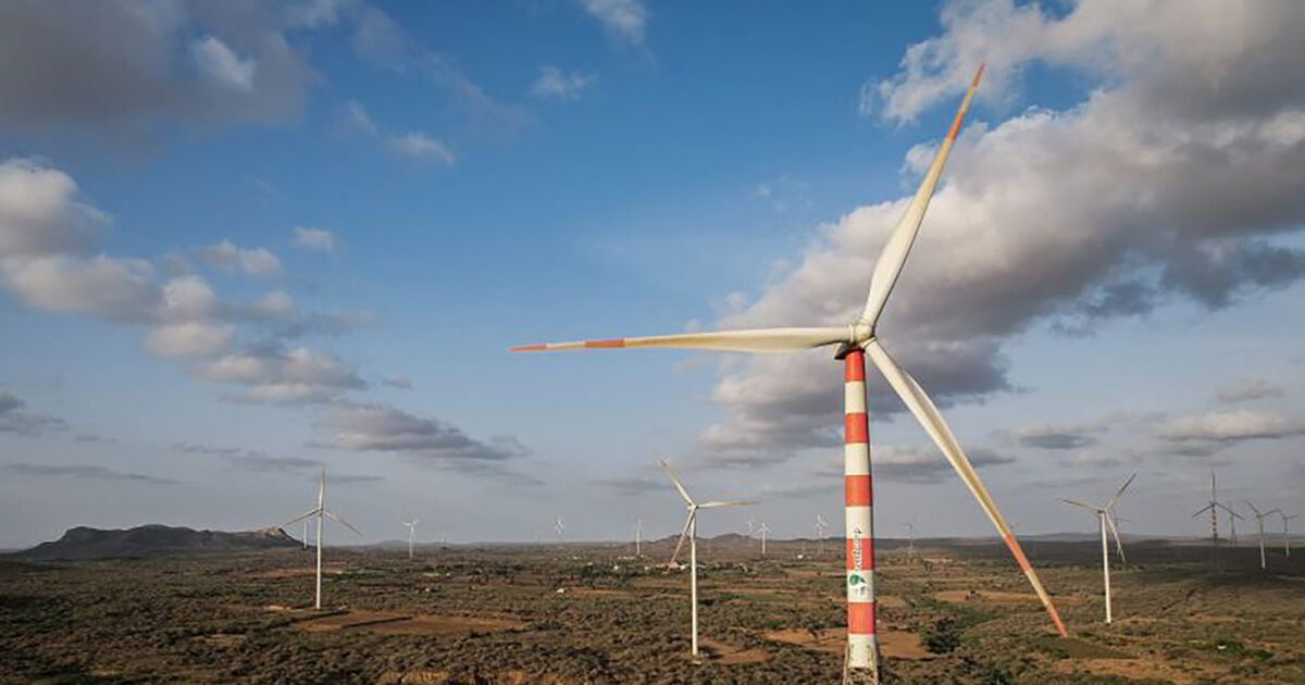 Vindkraftverk med röda och vita ränder står i ett öppet landskap under en delvis molnig himmel, med kullar och spridd vegetation i bakgrunden.