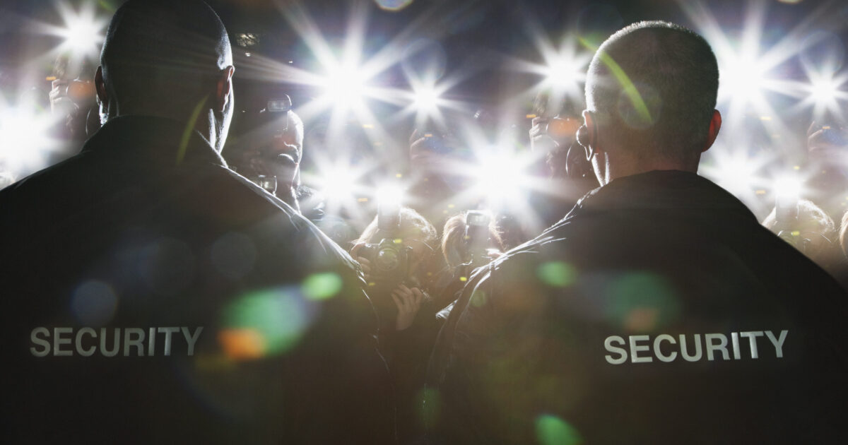 Två säkerhetsvakter står med ryggarna mot kameran, vända mot en folkmassa av fotografer och blinkande kameror i en ljus, hektisk scen.