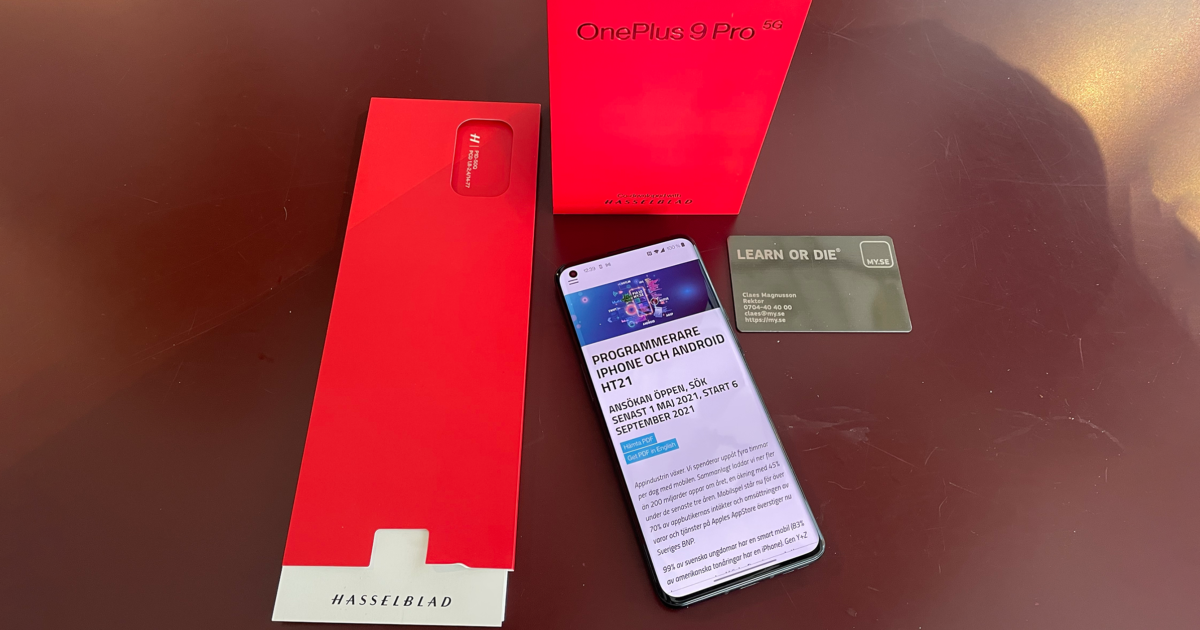 En OnePlus 9 Pro 5G-telefon med en webbplats ligger på en röd yta, bredvid sin röda låda, ett fodral med Hasselblad-märket och ett grått kreditkort märkt "LEARN OR DIE".