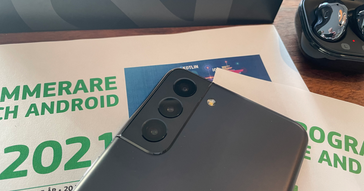 En närbild av en svart smartphone, med fokus på dess trippelkamera, placerad ovanpå papper med grön text och bredvid svarta trådlösa hörlurar i deras fodral.