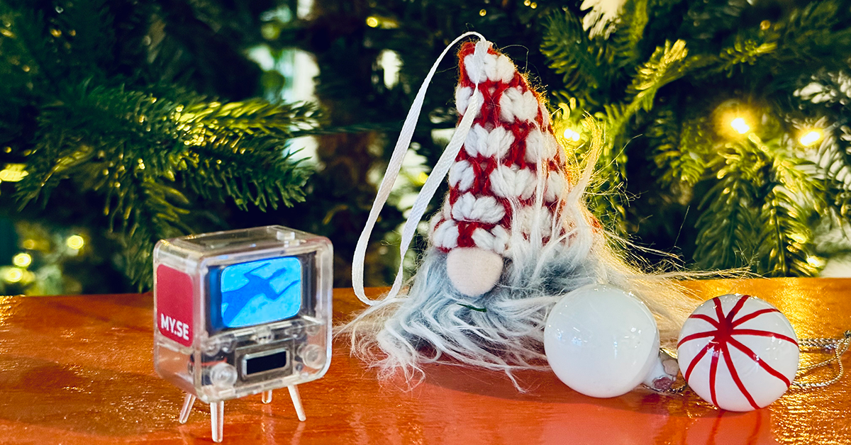 En liten leksaks-TV, en tomte-dekoration med en röd och vit hatt, en vit julgranskulorid och en vit julgranskulorid med röda ränder står på en orange yta framför en upplyst julgran.