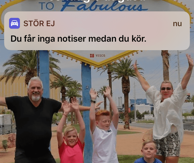 En familj på fem poserar exalterat framför en färgglad "Välkommen till Fabulous"-skylt med palmer; en telefonavisering säger "Du får inga aviseringar medan du kör".