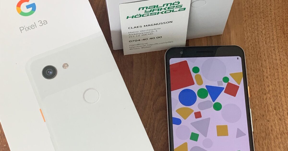 En Google Pixel 3a-smartphone ligger på en träyta bredvid sin låda och en annan Pixel 3a-låda. Ett visitkort från Malmö högskola ligger ovanpå en låda. Telefonens skärm visar ett färgglatt abstrakt mönster.