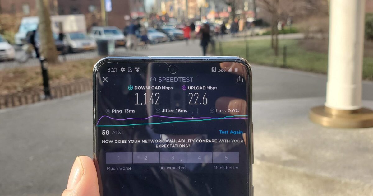 En hand håller en smartphone utomhus och visar ett hastighetstestresultat: 114,2 Mbps nedladdningshastighet och 22,6 Mbps uppladdningshastighet, med en livlig stadsgata och suddiga människor i bakgrunden.