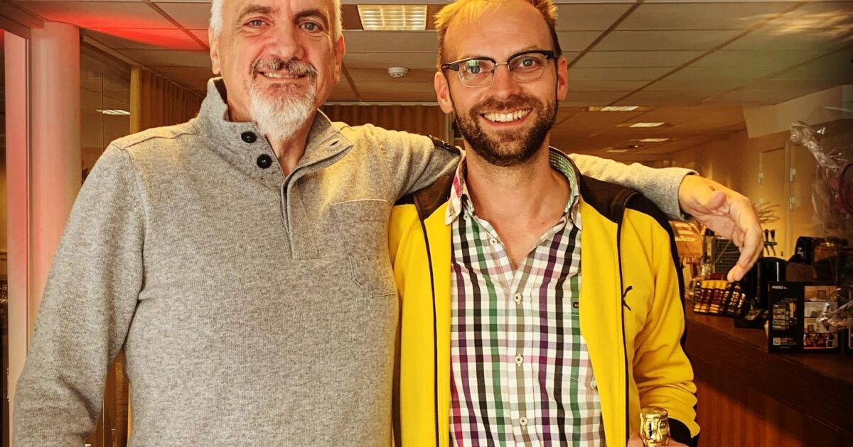 Två leende män står inomhus; mannen till vänster bär en ljusgrå tröja, medan mannen till höger, i gul jacka och rutig skjorta, håller en flaska. Mannen till vänster har armen om den andres axlar.