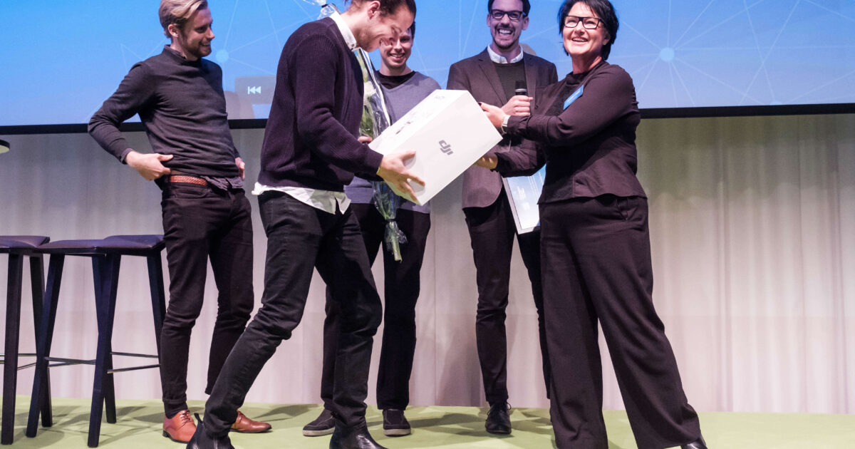 Fem personer står på en scen; en tar emot en stor vit låda och blommor från en leende kvinna, medan de andra tittar och ler. Det finns två pallar och en projektionsduk i bakgrunden.