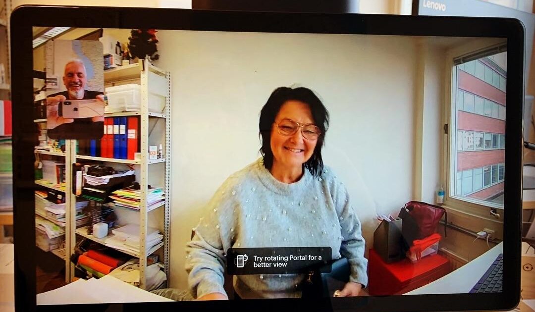 En kvinna ler under ett videosamtal, syns på en skärm på ett kontor. En man dyker upp i ett litet fönster i det övre vänstra hörnet. Kontoret har hyllor med filer, ett fönster och meddelandet Försök att rotera portalen för en bättre vy visas.