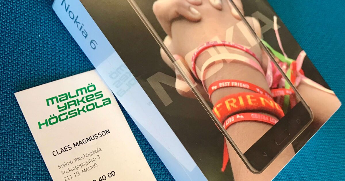 En låda med en Nokia 6-smarttelefon med två händer knäppta, tillsammans med ett visitkort från Malmö Yrkeshögskola för Claes Magnusson, placerat på en blå yta.