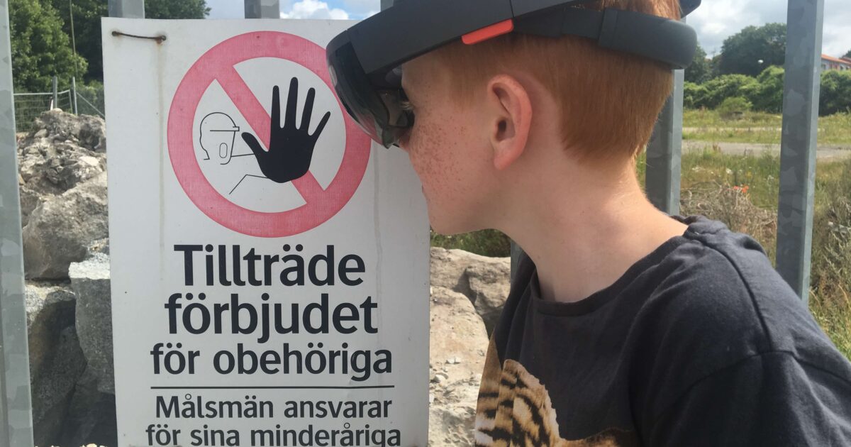 En pojke med VR-glasögon står nära ett metallstaket med en svensk varningsskylt som förbjuder obehörig åtkomst. Stora stenar syns bakom staketet och himlen är delvis molnig.