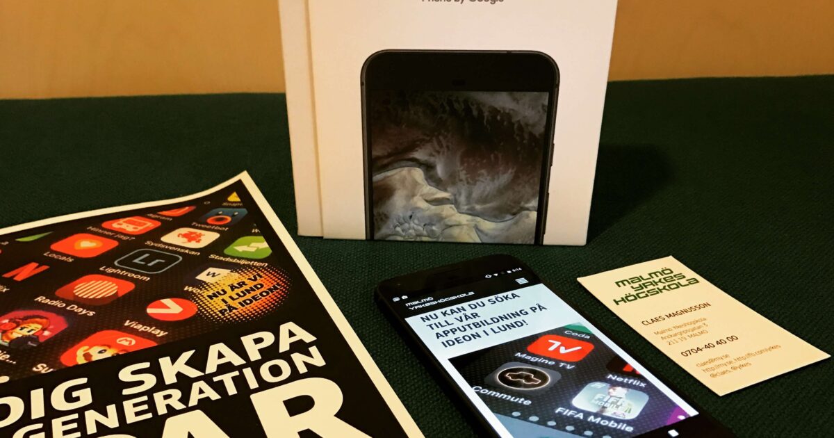 En Google Pixel-telefon, dess låda, en broschyr från Malmö Yrkeshögskola och ett visitkort visas på en mörkgrön yta. Telefonskärmen visar olika appikoner och svensk text.
