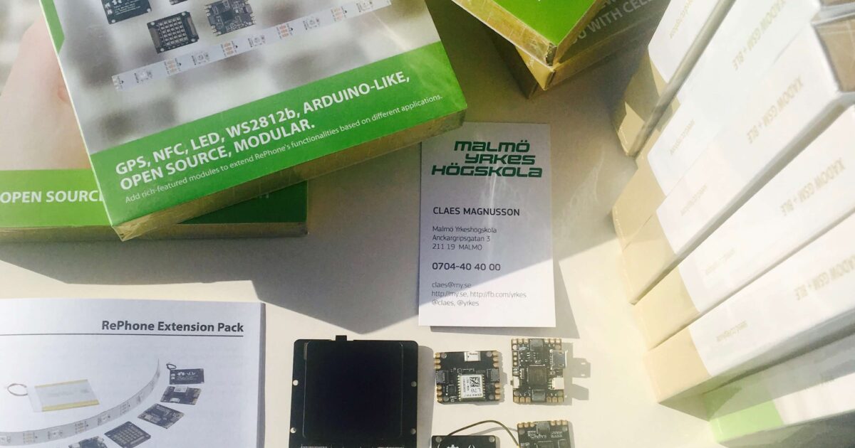 Blandade elektroniska moduler, kit, instruktionsblad och lådor från RePhone ligger utspridda på ett bord tillsammans med ett visitkort och tilläggspaket från Malmö Högskola, under stark belysning.