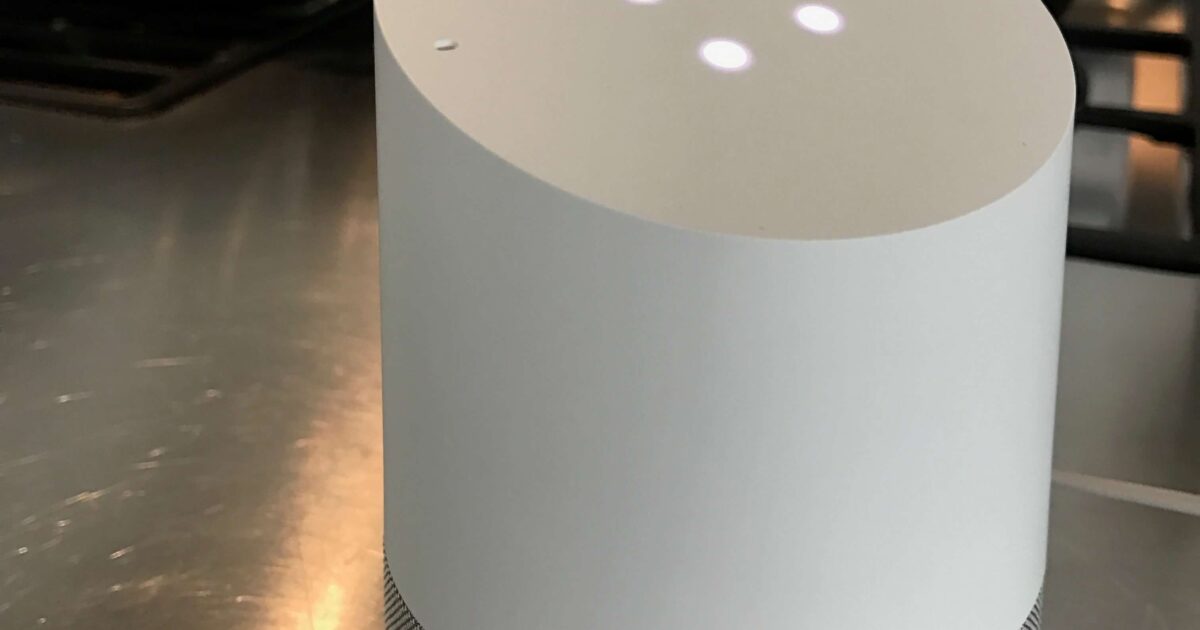 En vit smart högtalare med fyra lysande prickar på toppen står på en köksbänk i rostfritt stål nära en spis med en stekpanna och köksredskap i bakgrunden.