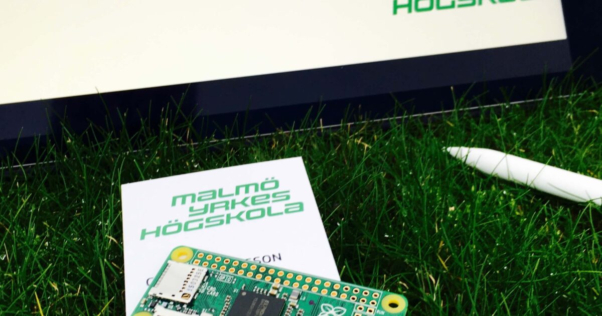 Ett Raspberry Pi-kort vilar på ett visitkort från Malmö Yrkeshögskola på gräs, med en penna och ett schematiskt diagram på en surfplatta i bakgrunden.