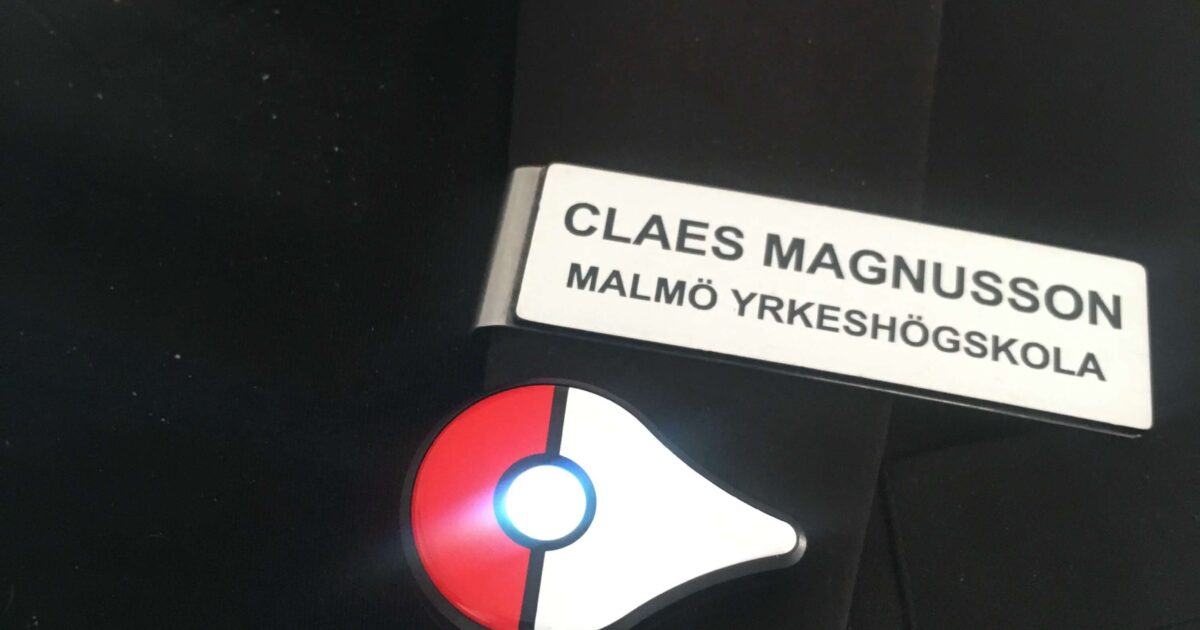 En person klädd i en svart jacka med en namnbricka där det står CLAES MAGNUSSON MALMÖ YRKESHÖGSKOLA och en Pokémon Go Plus-enhet med ett lysande blått ljus fäst under namnbricka.