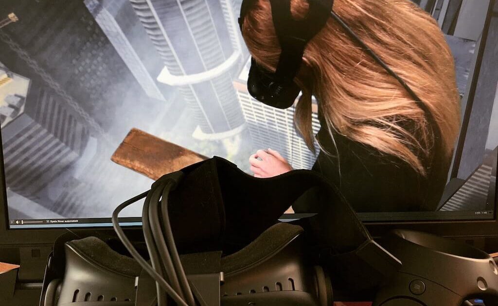 En person med VR-headset syns på en skärm och ser ut att titta ner från en hög byggnad i en virtuell stad. Framför den sitter ett HTC Vive-headset och handkontroller placerade på ett skrivbord.