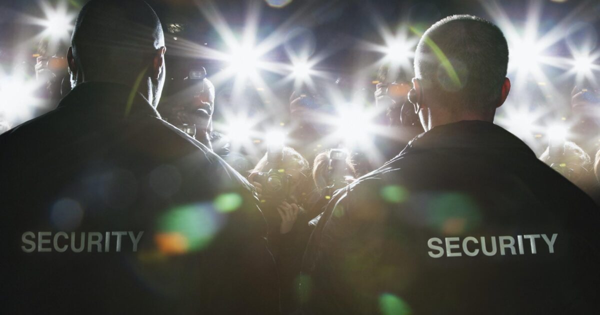 Två säkerhetsvakter står mitt emot en folkmassa av blinkande kameror och fotografer, med SÄKERHET tryckt på baksidan av deras jackor, upplysta av ljusa kamerablixtar.