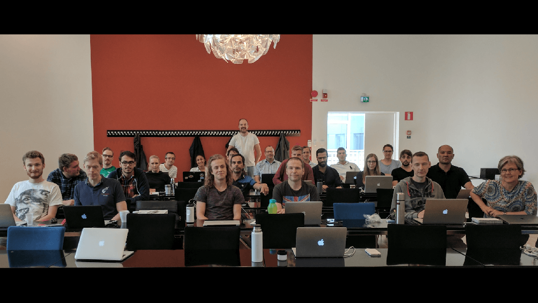 En grupp människor sitter vid skrivbord med öppna bärbara datorer i ett klassrum, vända mot kameran. En man står längst bak i rummet. Väggen bakom dem är röd, och ovanför finns en modern lampa.