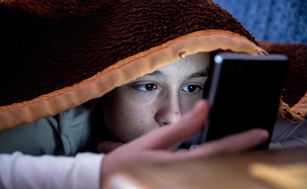 Ett barn ligger under en brun filt och tittar noga på en smartphone-skärm, med ansiktet upplyst av enhetens ljus.