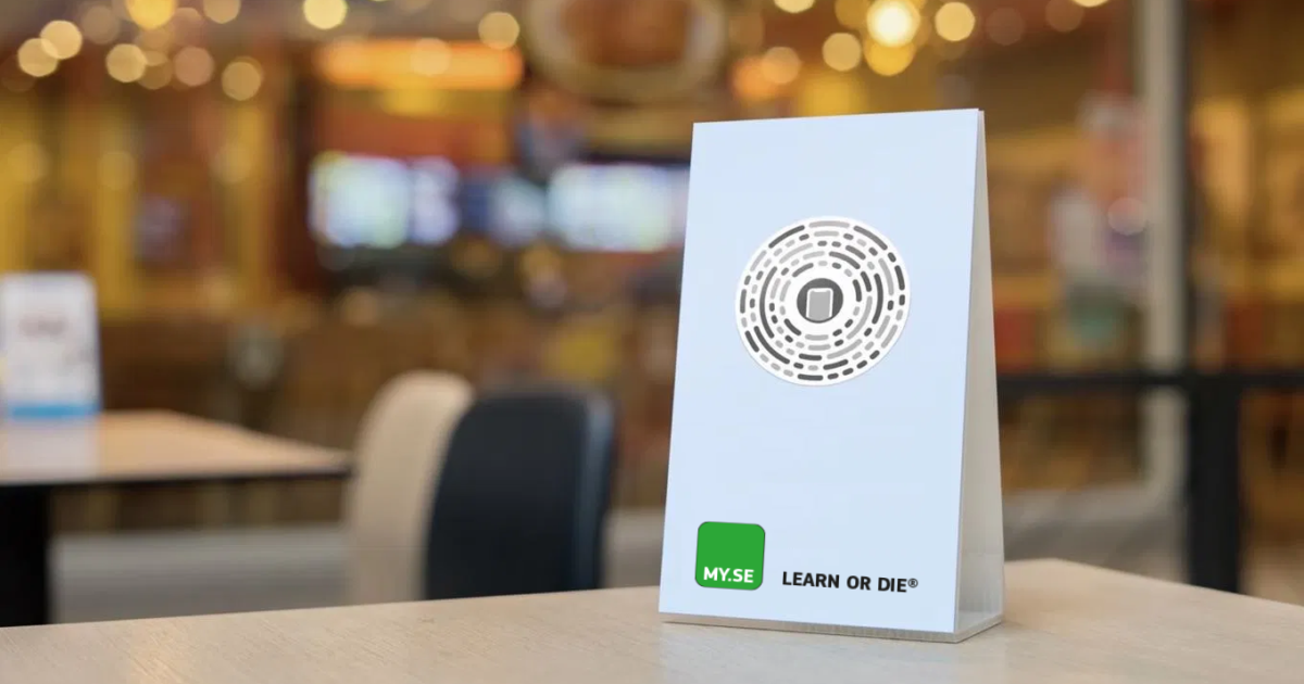 En vit bordsskylt med cirkulär design och texten MYSE LEARN OR DIE® står på ett ljust bord i en suddig kafé- eller restaurangmiljö.