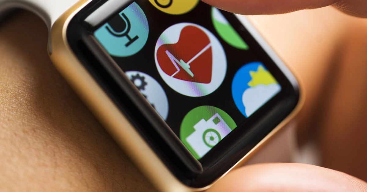 En närbild av en smartklockas skärm som visar appikoner, med ett finger som är på väg att trycka på en hjärtformad hälso- eller pulsapp. Smartklockan sitter på en persons handled.