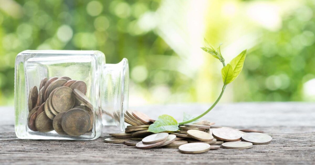 En glasburk vält över på en träyta, mynt spills, med en liten grön växt som spirar ur mynthögen. Bakgrunden är suddig grönska.