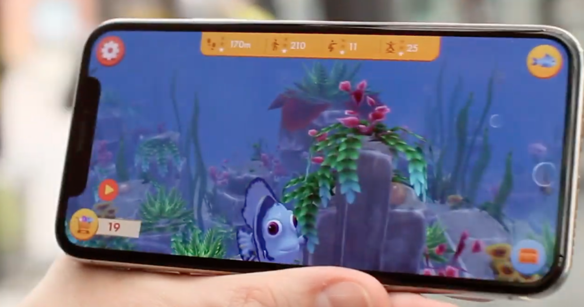 En hand håller en smartphone som visar ett spel med undervattenstema, tecknade fiskar, färgglada växter och spelstatistik längst upp på skärmen.