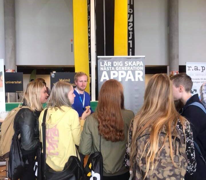 En grupp ungdomar med ryggsäckar står framför en monter om appskapande och lyssnar på en representant. Gula pelare och informationsbanderoller syns i bakgrunden.