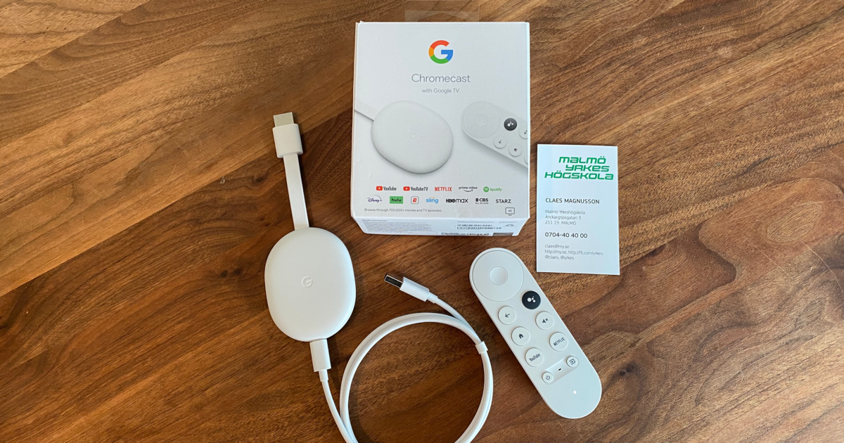En Chromecast med Google TV-enhet, fjärrkontroll, produktförpackning och ett visitkort visas på en träyta. Chromecast och fjärrkontroll är vita; förpackningen visar Googles logotyp och streamingtjänsternas logotyper.