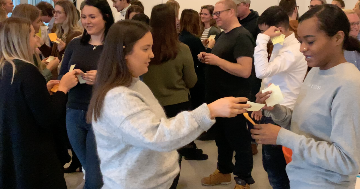 En grupp människor står och interagerar i ett starkt upplyst rum, utbyter post-it-lappar och pratar i små grupper, vilket föreslår en samarbets- eller nätverksaktivitet.