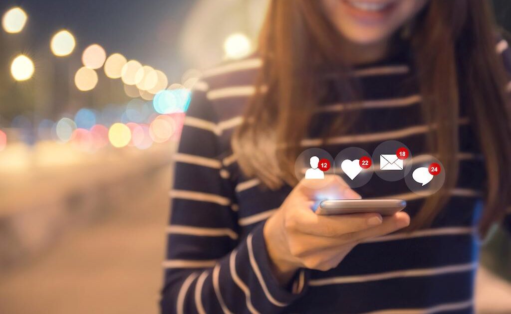 En kvinna i randig skjorta ler medan hon använder sin smartphone på natten, med ikoner för sociala medier-aviseringar, inklusive gilla-markeringar, meddelanden och följare, som svävar ovanför telefonen.
