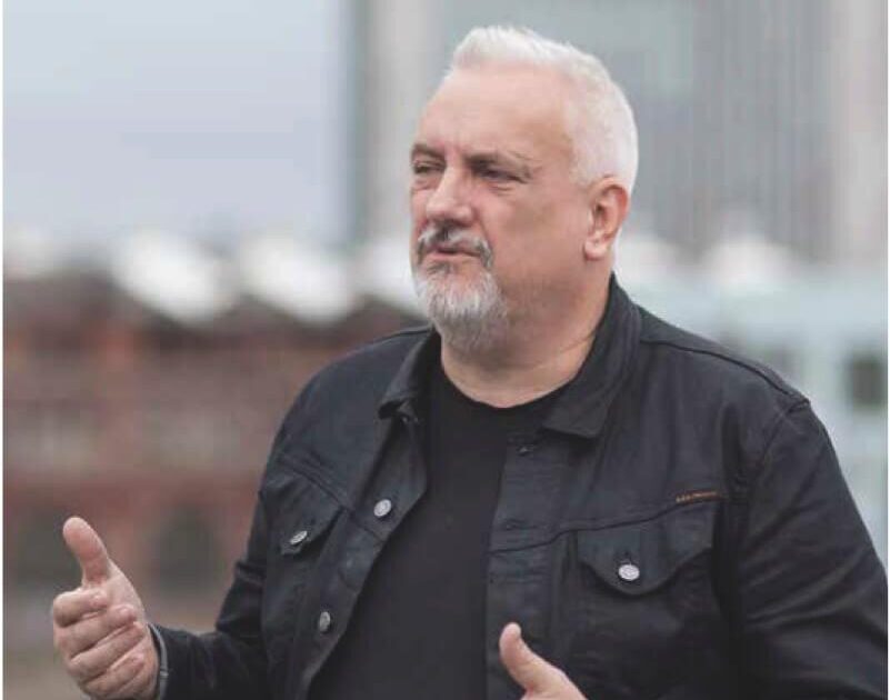 En medelålders man med kort grått hår och skägg står utomhus och gestikulerar med händerna. Han bär en svart jacka över en mörk skjorta, med en suddig stadsbild i bakgrunden.