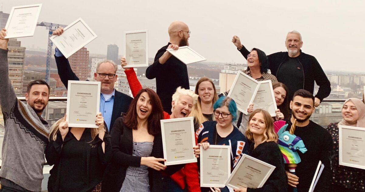 En grupp människor står på ett tak, håller i sina certifikat och ler upphetsat. Stadens silhuett syns i bakgrunden en molnig dag.