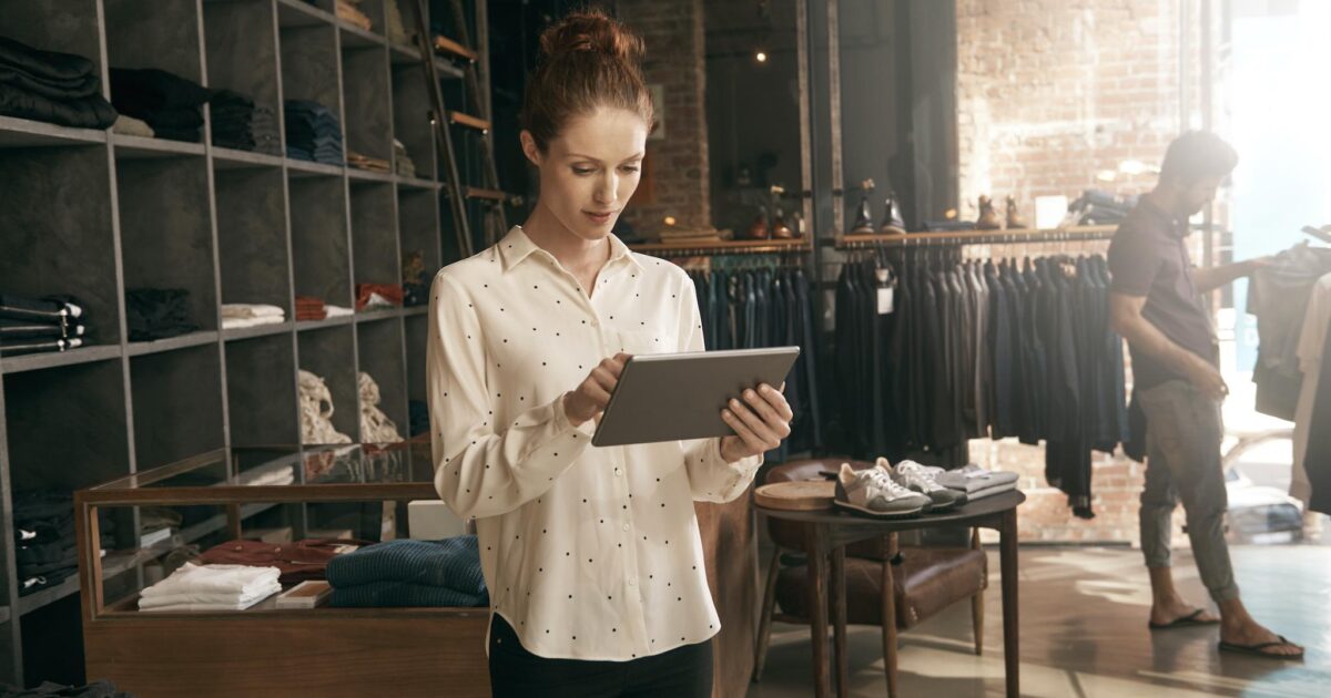 En kvinna i vit blus använder en surfplatta i en modern klädaffär, med hyllor med vikta kläder och hängande plagg i bakgrunden. En man tittar på varor nära fönstret. Varmt, naturligt ljus fyller utrymmet.