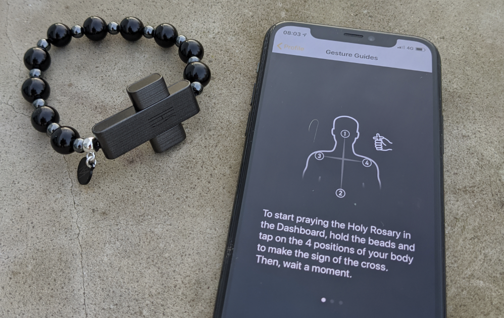 Ett pärlarmband med ett korsberlock ligger bredvid en smartphone som visar instruktioner för att göra korstecknet som en del av att be rosenkransen med hjälp av en app.