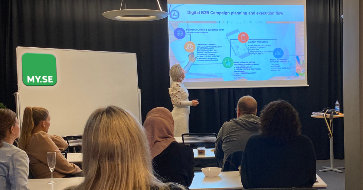 En kvinna presenterar en bild med en digital B2B-kampanjplanering för en sittande publik i ett modernt klassrum, med en whiteboardtavla och en stor MY.SE-logotyp synlig. Publiken lyssnar uppmärksamt, och några antecknar.