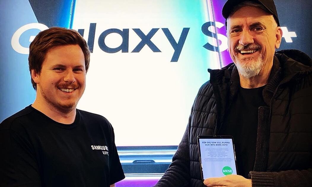 Två leende män står framför en stor Samsung Galaxy S9+-skärm. Den ena bär en Samsung Expert-tröja, medan den andra, i svart keps och jacka, håller en plakett eller ett certifikat med båda händerna.