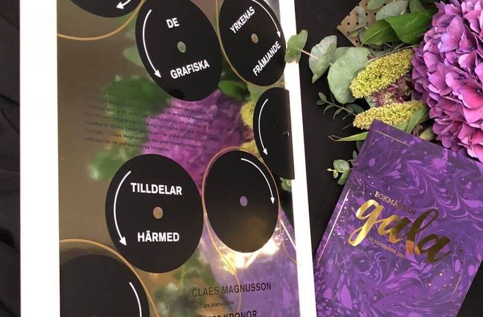 En inramad utmärkelse med svarta cirkulära plaketter och svensk text bredvid en lila blomsterbukett och ett lila marmorerat häfte med guldskrift på en mörk yta.
