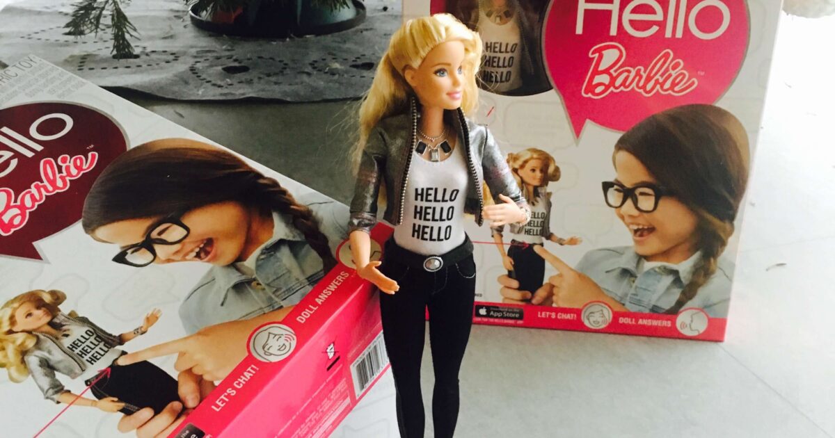 En Hello Barbie-docka står framför sin förpackning under en julgran. Lådan har bilder av dockan, och ett litet kort ligger på golvet i närheten.