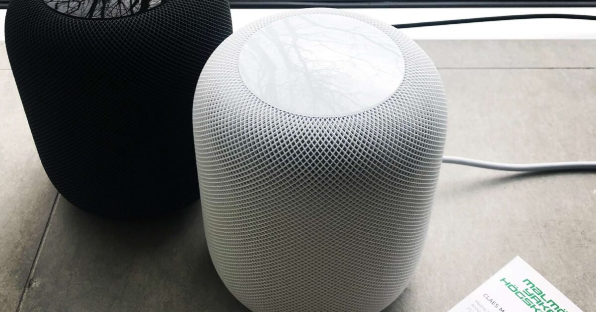 En svart och en vit smarthögtalare står sida vid sida på ett grått golv nära ett fönster, med ett visitkort från ett teknikföretag bredvid dem. Gräs syns utanför genom glaset.