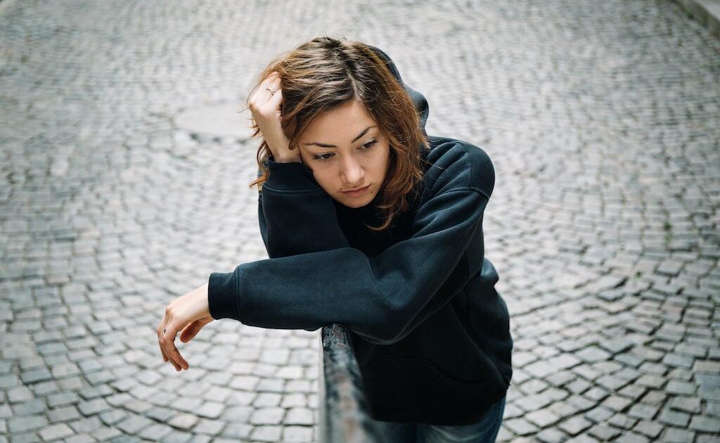 En ung kvinna i svart lutad huvtröja lutar sig mot ett metallräcke och tittar fundersamt ner, med en kullerstensgata i bakgrunden.