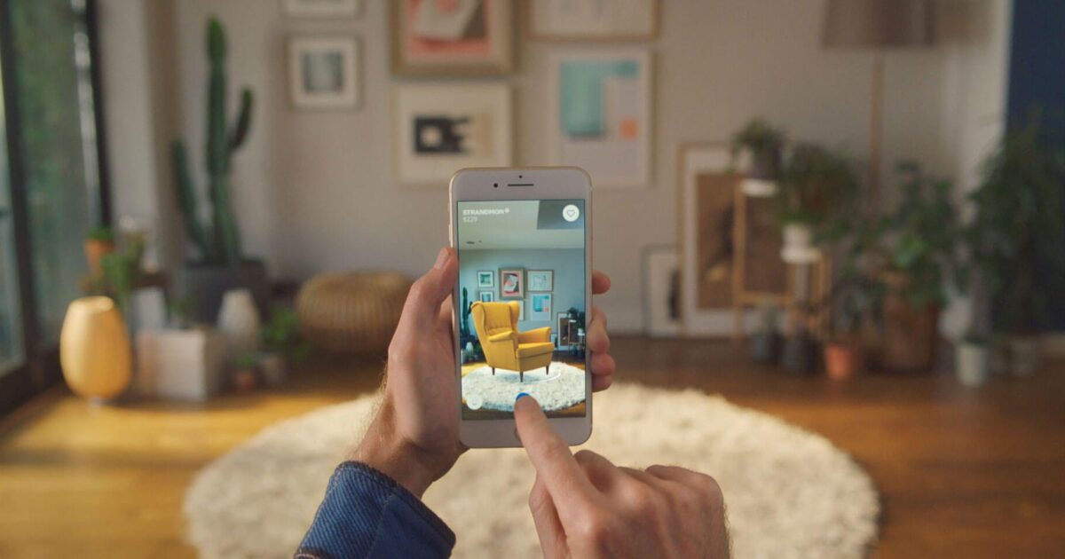En person använder en smartphone för att visualisera en gul fåtölj i sitt vardagsrum med en app för förstärkt verklighet; det riktiga rummet har växter, inramad konst och en rund matta.