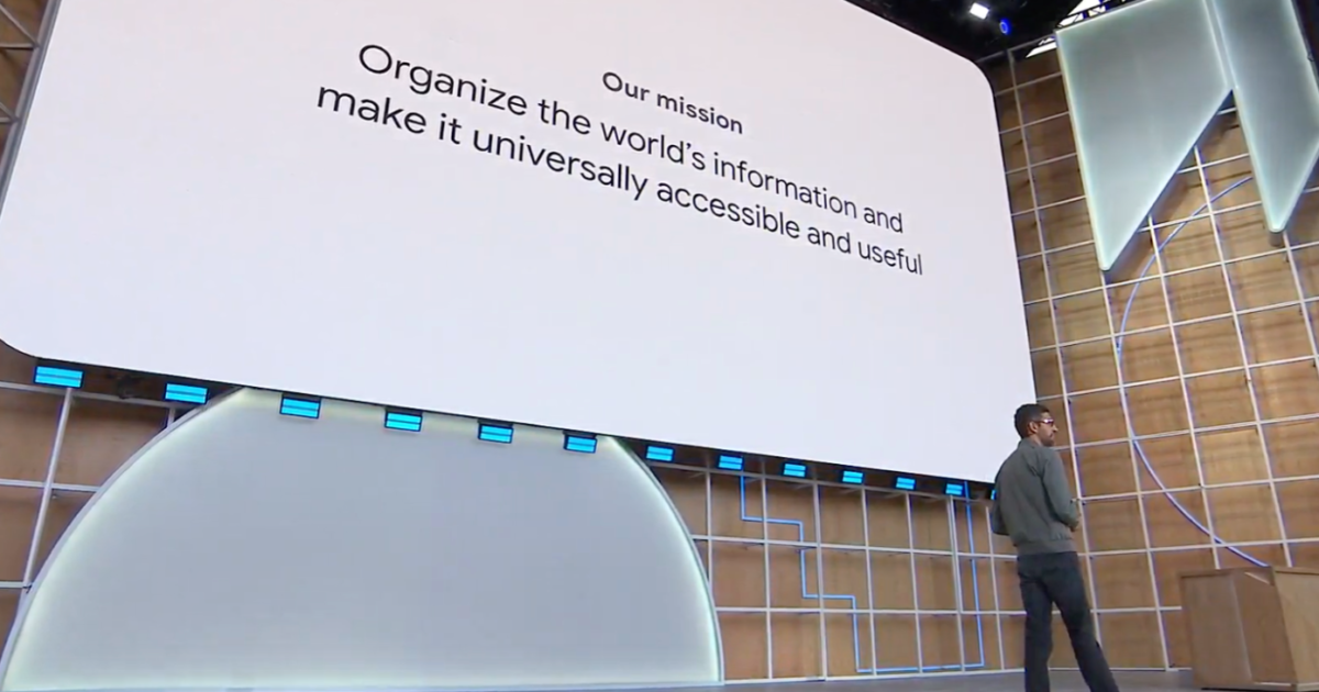 En person står på scenen framför en stor skärm och visar texten: Vårt uppdrag: Organisera världens information och göra den universellt tillgänglig och användbar. Miljön ser ut att vara en teknikkonferens eller presentation.