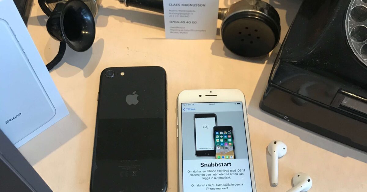 En modern iPhone, AirPods och en iPhone-låda visas bredvid en gammal telefon med vridknapp och hörlur på ett skrivbord. Ett visitkort och en installationsskärm på iPhonen syns också.